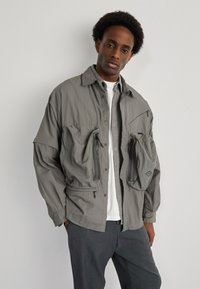 Gray lightweight jacket with two zippered pockets, button-up collar, and a relaxed fit. Worn over a white shirt and gray pants.