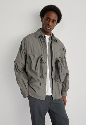 Gray lightweight jacket with two zippered pockets, button-up collar, and a relaxed fit. Worn over a white shirt and gray pants.
