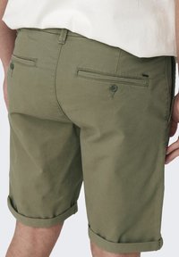 Shorts en coton vert olive avec une coupe droite, des poignets retroussés et deux poches arrière avec fermetures à bouton et des détails de coutures subtils.