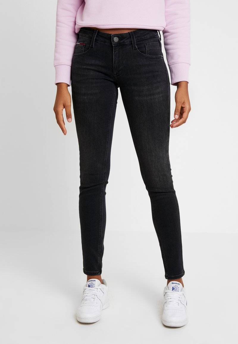 Jeans skinny negros de denim con un ligero estiramiento. Presentan un diseño clásico de cinco bolsillos y un lavado oscuro. Se combinan con zapatillas blancas.