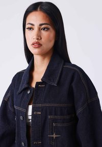 Giacca di jeans scuro con colletto a giro, ampie tasche frontali, cuciture visibili e dettagli metallici. Presenta una vestibilità rilassata e un tessuto testurizzato.