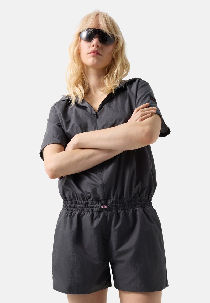 Lyshåret person iført oversized sorte solbriller og en sort kortærmet zip-op romper med elastisk talje, armene er krydset.