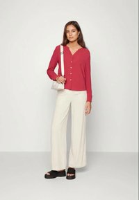 Camisa vermelha com botões, de mangas longas, calças largas bege e sandálias de plataforma pretas. Uma pequena mala de ombro em creme completa o visual.