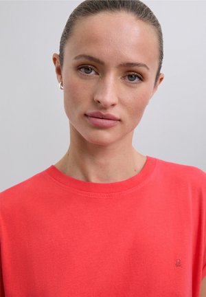 Jeune femme à la peau claire et aux yeux verts portant un t-shirt corail rouge, regardant calmement l'appareil photo sur un fond clair.
