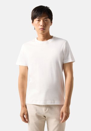 En ung mand står med fronten mod kameraet iført en ensfarvet hvid T-shirt med korte ærmer og lyse beige bukser mod en hvid baggrund.