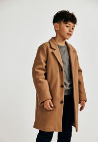 Kameli värvi villane mantel, millel on nurgeline krae, ühe nööbiga disain ja suured nööbid, kantud halli sviitri kohal.