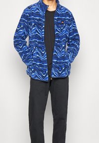 Giacca in pile blu con un motivo ondulato, dotata di un collo alto, zip centrale e tasche laterali. Abbinata a una maglietta nera e pantaloni.