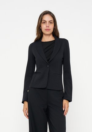 Femme en blazer noir à un seul bouton et pantalon noir assorti, debout devant un fond blanc uni, regardant droit devant avec une expression neutre.