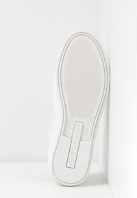 Semelle blanche de basket avec motif antidérapant texturé et logo "MICHAEL KORS" embossé au centre, positionnée verticalement contre un coin blanc.