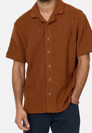 Uomo che indossa una camicia marrone a maniche corte, strutturata, con colletto e bottoni, indossata fuori sopra pantaloni blu scuro, una mano in tasca.