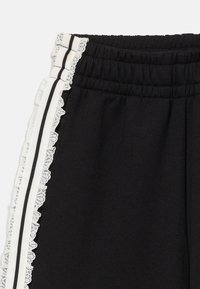 Schwarze Hose mit elastischem Bund, weißen Seitenstreifen und einer Spitzenrüsche entlang der äußeren Nähte.