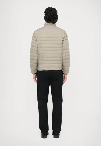 Odebana svetlo siva bunda z vodoravnimi šivi in visokim ovratnikom, kombinirana s črnimi hlačami in črnimi čevlji, prikazana od zadaj.