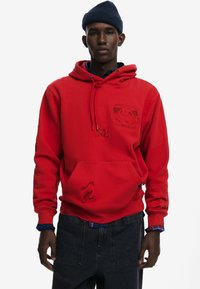 Červená mikina s kapucňou, predným vreckom a vyrazenými vzormi. Má rebrovaný lem a manžety. Nosená cez tmavé džíny s károvaným detailom.