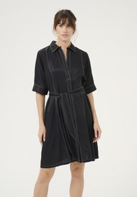 Melnas krekla kleitas ar īsajiem piedurknēm, pogu aizdare, jostas vieta, kontrastējoša balta dūriens un brīva piegriezuma. Gluda auduma tekstūra.