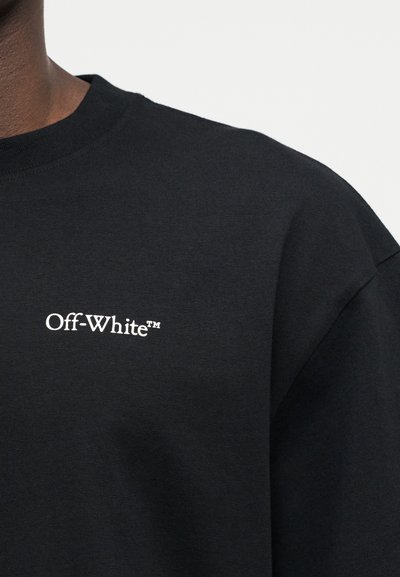 Μαύρο μπλουζάκι με στρογγυλή λαιμόκοψη που φέρει μικρό λευκό λογότυπο "Off-White™" αριστερά στο στήθος, φορεμένο από ένα άτομο μπροστά σε απλό φόντο.