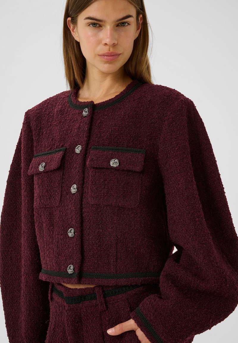 Bordeaux-farbene cropped Jacke aus strukturiertem Material, mit zwei aufgesetzten Taschen, silbernen Knöpfen und grünem Besatz am Halsausschnitt und Saum.