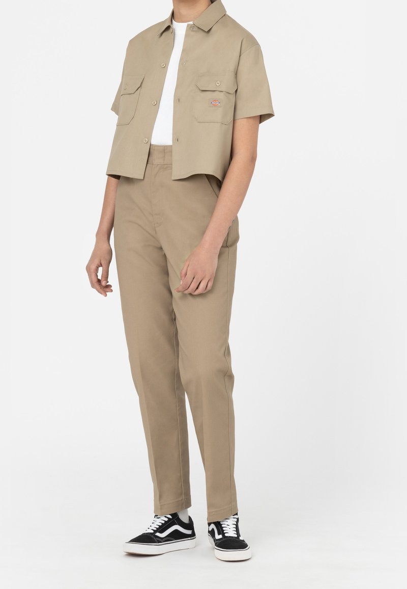 Personne portant une chemise beige à manches courtes boutonnée et un pantalon taille haute assorti avec des baskets noires et blanches sur un fond uni.