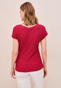 Camiseta roja de textura con corte holgado, mangas cortas y un escote amplio. La tela presenta un patrón arrugado y aberturas laterales.
