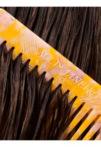 Peine para el cabello con patrón amarillo y rosa etiquetado como "Sol de Janeiro", peinando mechones de cabello oscuro, húmedo y liso contra un fondo oscuro.