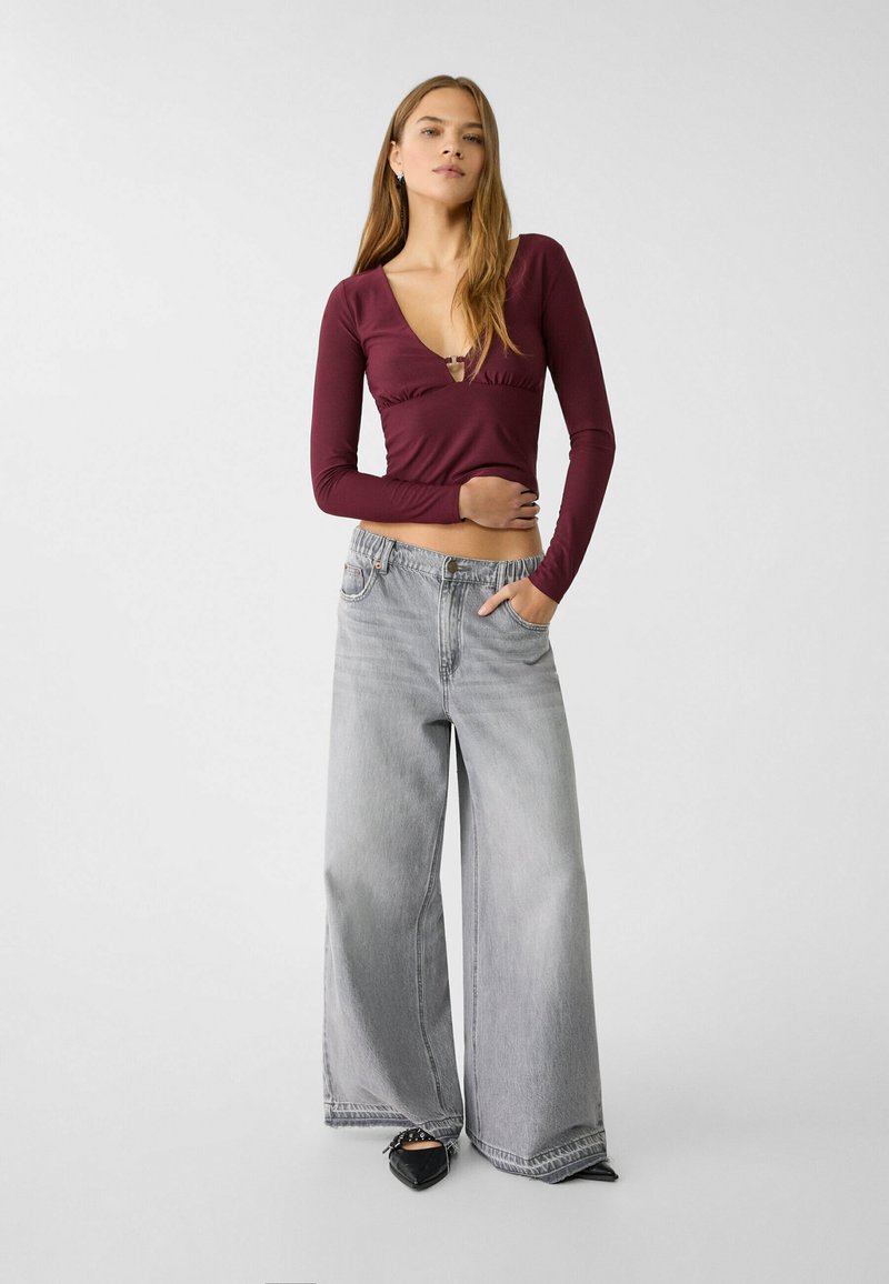 Maglione lungo bordeaux con scollatura a V profonda e dettagli arricciati, abbinato a jeans grigi chiari a gamba larga e ballerine nere con punta affusolata.