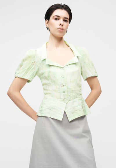 Πράσινο πουκάμισο μέντας με κοντό μανίκι, με δομημένο γιακά, ζώνη πεπλούμ και έξι κουμπιά μπροστά. Ομαλή υφή με διακριτικό μαρμαροειδές μοτίβο.