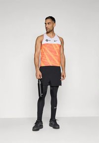 Sporta apģērbs ar spilgti oranžu un baltu bezroku krekliņu, melniem šortiem, melnām legingiem un melniem sporta apaviem, turot melnu ūdens pudeli.