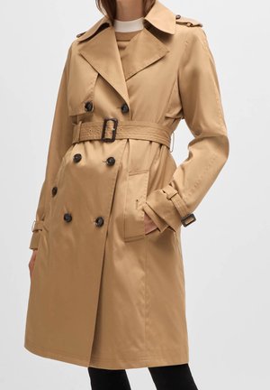 Vrouw in een beige dubbele trenchcoat met zwarte knopen, een tailleband en banddetails op de mouwen, staand met één hand in de zak.