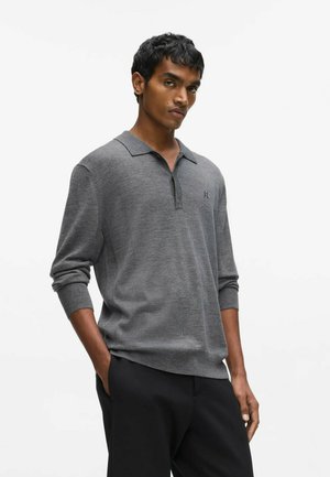 Young man with short dark hair wearing a gray long-sleeve polo shirt and black pants, standing with one hand in pocket against white background.