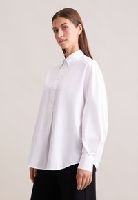 Camicia bianca con colletto, maniche lunghe e vestibilità comoda. Presenta una placca frontale e un tessuto leggermente strutturato.