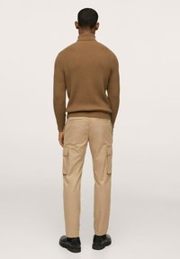 Pull à col roulé côtelé marron, pantalon cargo beige avec poches latérales, et chaussures noires, vus de dos sur un fond neutre.