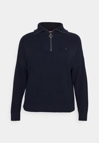 Granatowy sweter dzianinowy z długimi rękawami, zamek błyskawiczny na ćwierć długości w kolorze srebrnym oraz małym czerwonym, białym i niebieskim logo na piersi.