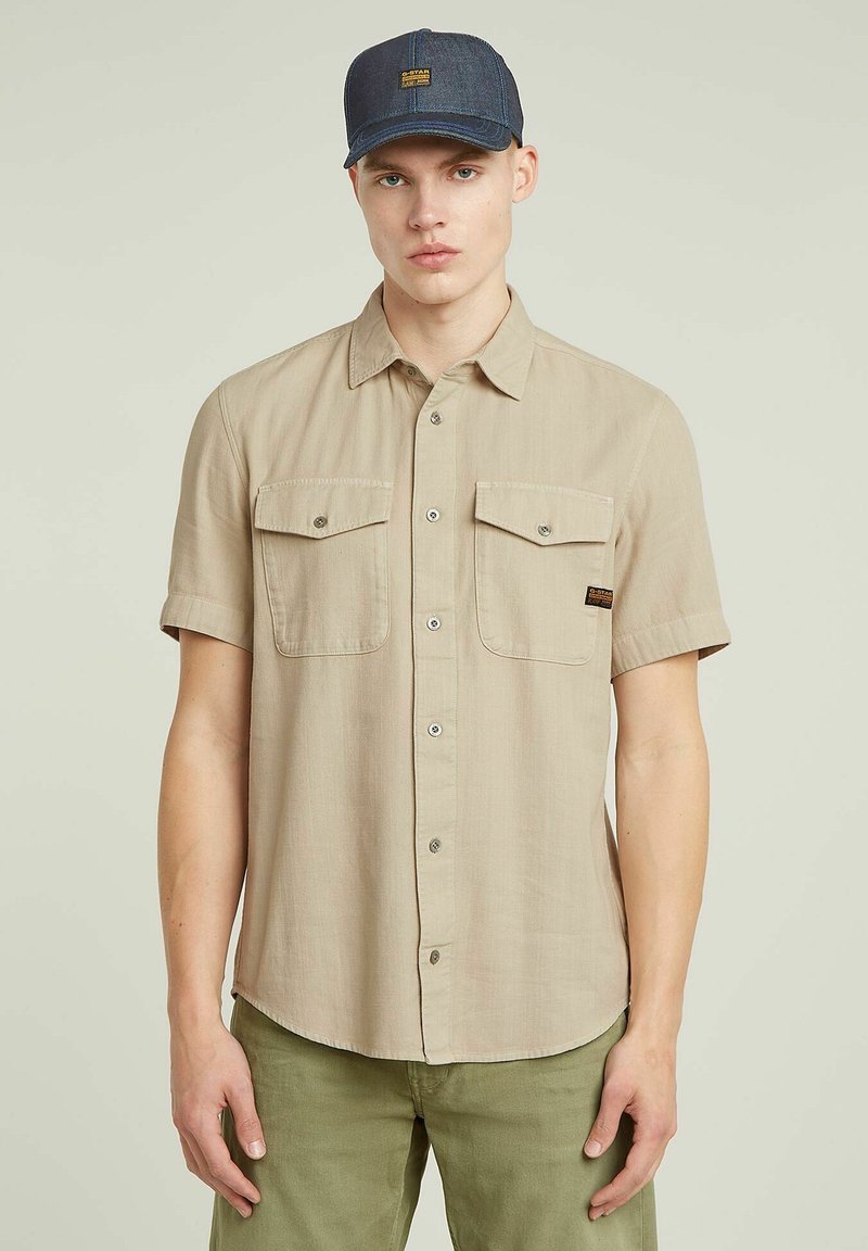 Beige overhemd met korte mouwen en een knoopsluiting aan de voorkant, twee borstzakken en zilveren knopen. Draagt een blauwe denim pet en groene broek.
