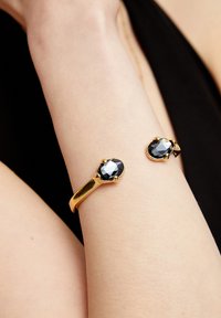 Pulsera de oro con dos piedras negras facetadas, acabado liso, forma ovalada y cierre seguro, lucida en un brazo desnudo.