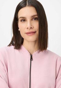 Lichtroze zip-up hoodie met een hoge halslijn, zachte stof en zwarte ritsdetail. Glad textuur, geen patronen of accenten zichtbaar.