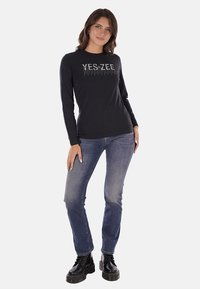 Maglietta nera a maniche lunghe con testo "YES ZEE" in argento, indossata con jeans blu e stivali neri stringati. Design semplice, vestibilità casual.
