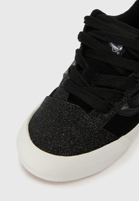 Musta urheilukenkä, jossa on kimalteleva pinta, mokka yksityiskohtia, mustat nauhat ja valkoinen kumipohja. Huomattava logo kielen kohdalla.