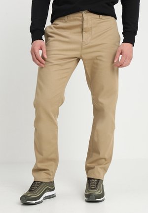 Mann trägt beige Hose, schwarzes Langarmshirt und olivgrüne Sneaker, steht vor einem schlichten hellen Hintergrund.