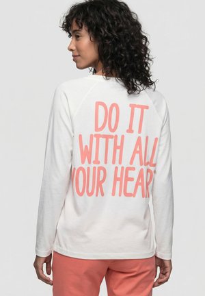 Woman with curly hair wearing a white long-sleeve shirt with coral text "DO IT WITH ALL YOUR HEART" and coral pants, facing sideways.