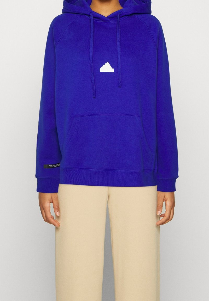 Sweat à capuche bleu avec un logo triangulaire blanc sur la poitrine, poche kangourou et poignets côtelés. Associé à un pantalon beige ample.