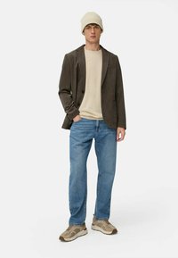 Mørkebrun kordfløjl blazer over en beige skjorte, parret med blå jeans og beige sneakers. Modellen bærer en lys beige beanie.