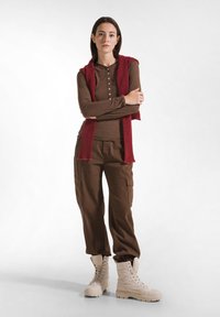 Bruine lange mouwen top met knopen, gecombineerd met bruine cargobroeken. Rode gestructureerde sjaal over de schouders gedrapeerd. Witte veterschoenen.