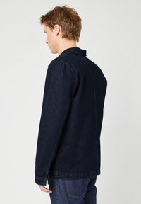 Giacca in denim blu scuro con colletto classico, maniche lunghe e taglio dritto. Presenta cuciture robuste e una finitura testurizzata.