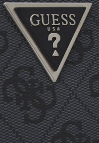 Svart texturerat material med en triangulär silverlogo som har "GUESS USA" och en utformning av ett frågetecken. En subtil mönstring synlig i bakgrunden.