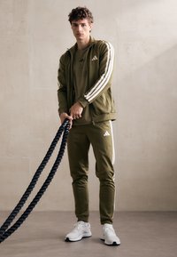 Olíva zöld sportos tréningruha, fehér csíkokkal, zipzáros kabáttal és testhezálló nadrággal, fehér sportcipővel párosítva. Fekete kötelet tart.