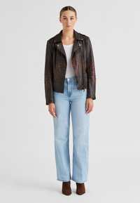 Braune Lederbikerjacke mit Leopardenmuster-Akzenten, Reißverschluss vorne und Revers. Kombiniert mit hellblauen weit geschnittenen Jeans und braunen Stiefeletten.