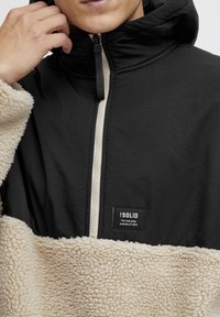 Pullover in pile nero e crema con zip frontale, tessuto testurizzato, cappuccio e un'etichetta sul lato sinistro. La parte superiore è elegante e liscia.