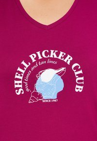 Camiseta de algodón fucsia con escote en V que presenta un gráfico de concha en blanco y azul claro y el texto: "CLUB DE RECOLECTORES DE CONCHAS, buenos momentos y líneas bronceadas, DESDE 1987."