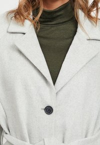 Lys grå uldblandingsfrakke med klapkrave, enkelt knaplukning og bælte, lagd ovenpå en mørkegrøn turtleneck.