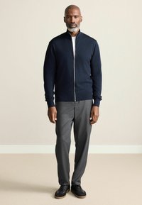 Marineblå zip-sweater med høj krave, parret med grå dressbukser og sorte sneakers. Glat tekstur, tætsiddende design og neutrale farver.