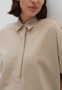 Camicia beige con colletto e una chiusura a bottoni frontale, realizzata in tessuto morbido, con maniche corte e una vestibilità morbida.